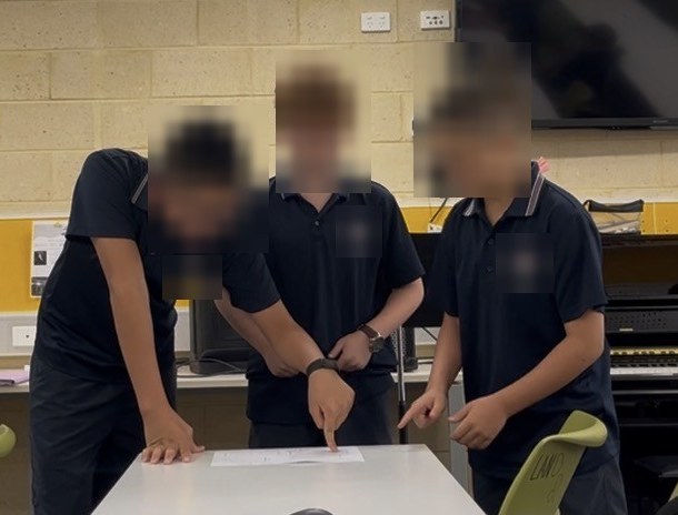 Three students standing around a desk, pointing at a rhythm sheet